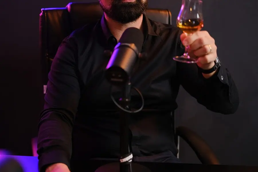 Apresentador sentado à mesa do Estúdio Mesacast, com microfone, taça e neon ao fundo