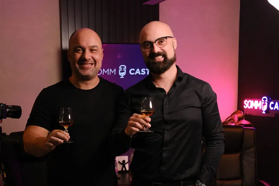 Dois apresentadores em pé no Estúdio Mesacast, diante do letreiro em neon