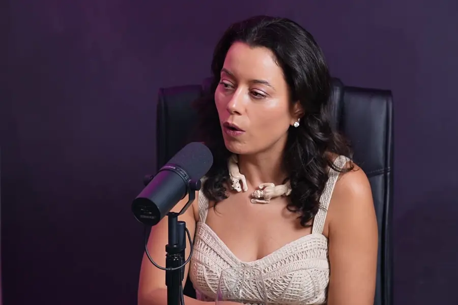 Apresentadora falando ao microfone, com parede violeta ao fundo