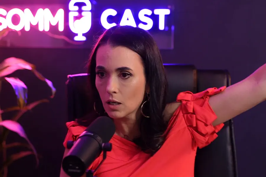 Convidada de vermelho falando ao microfone, com letreiro em neon e luz magenta ao fundo
